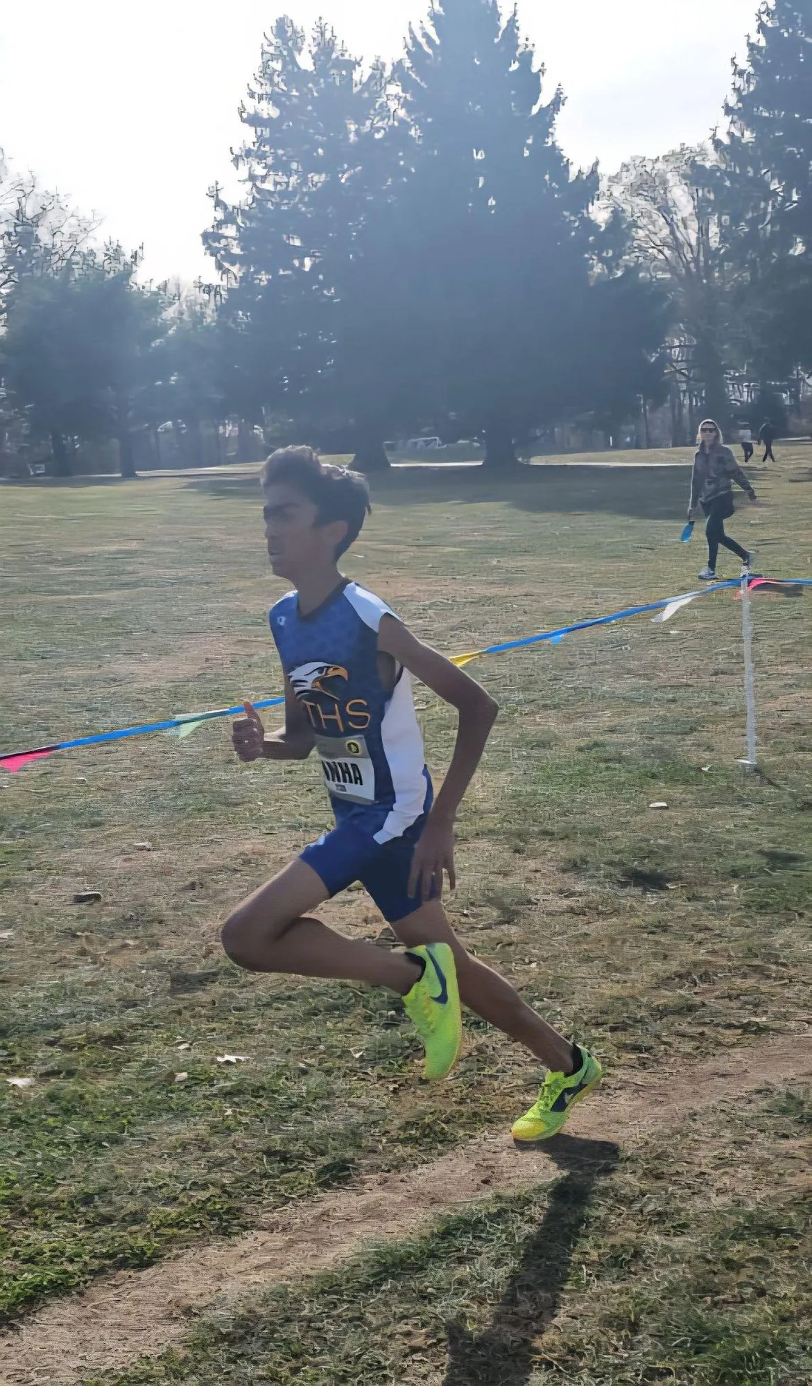 Anish mid-stride in a race on a grass course, wearing a blue singlet and yellow spikes