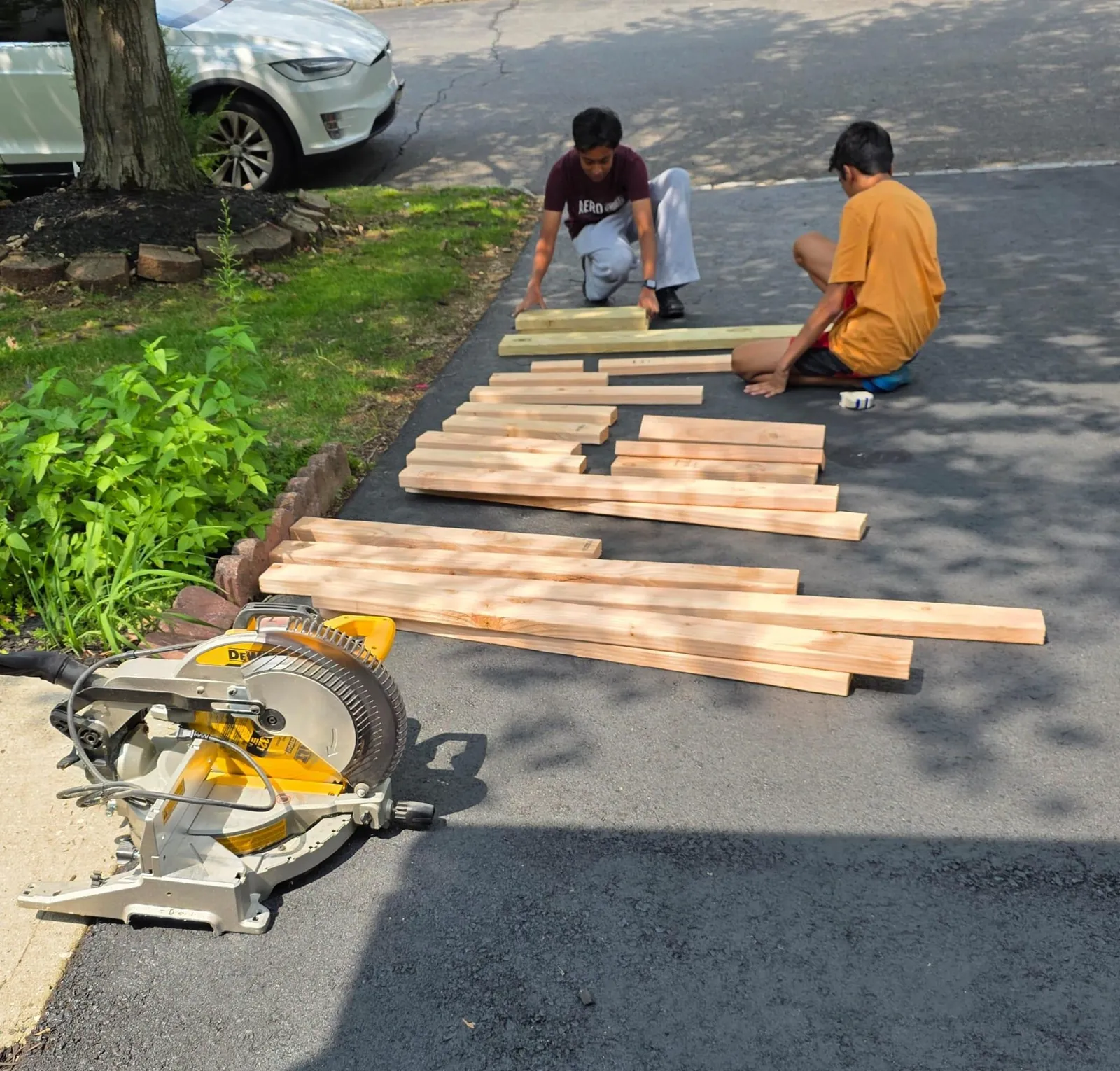 Cut lumber laid out on a driveway next to a miter saw, two people sorting pieces