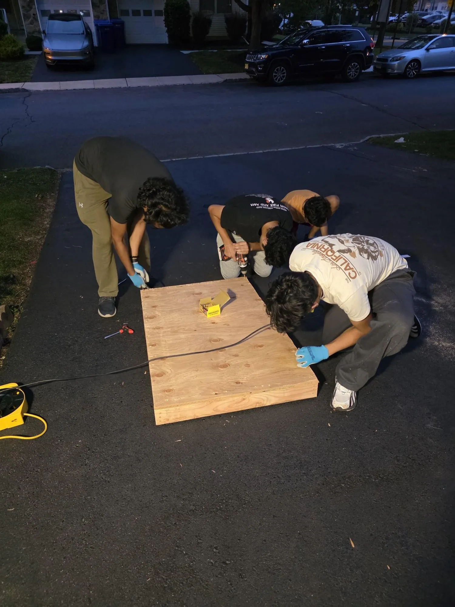 Four people working on the plywood floor pan on a driveway at night