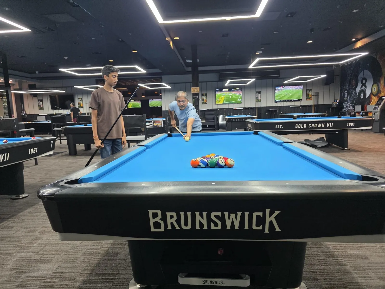 Anish holding a pool cue next to a blue billiards table while someone breaks