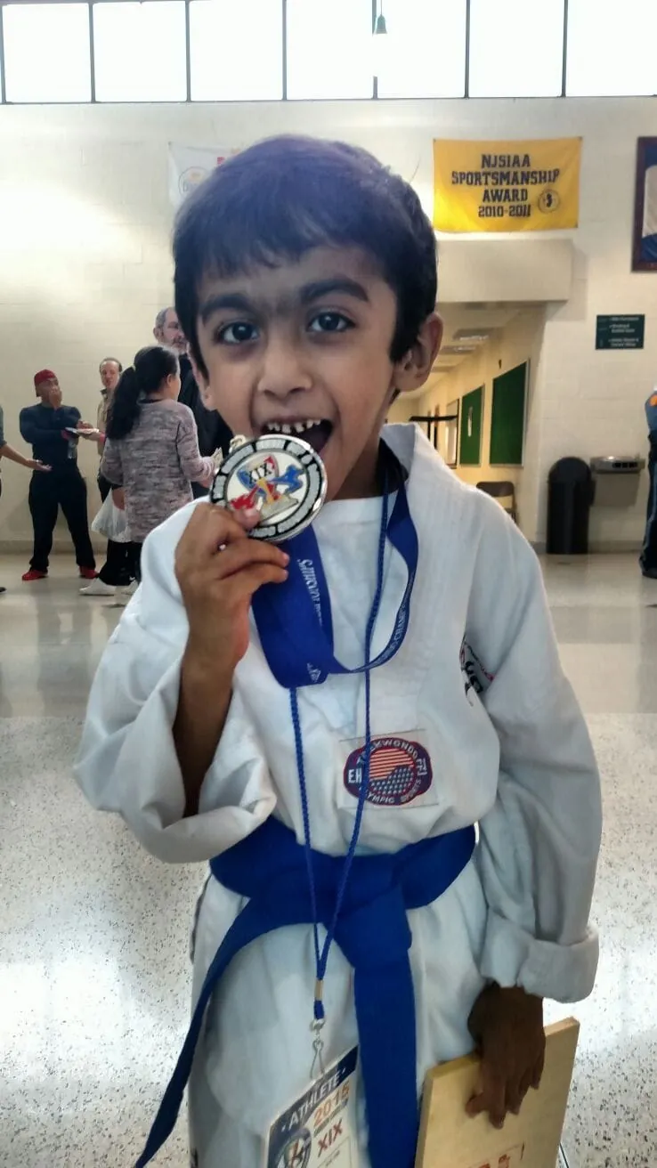 A young Anish in a taekwondo uniform with a blue belt, biting a tournament medal