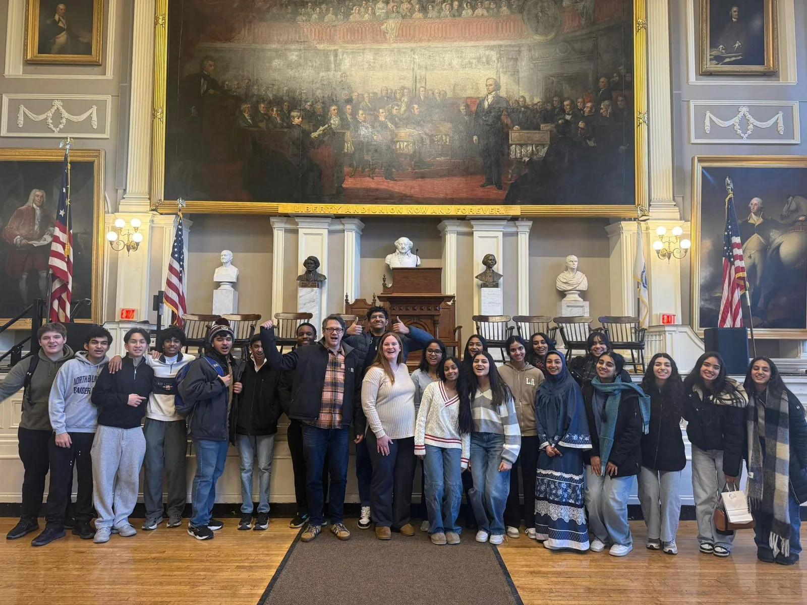 Large group of students posing in a historic hall with paintings and busts